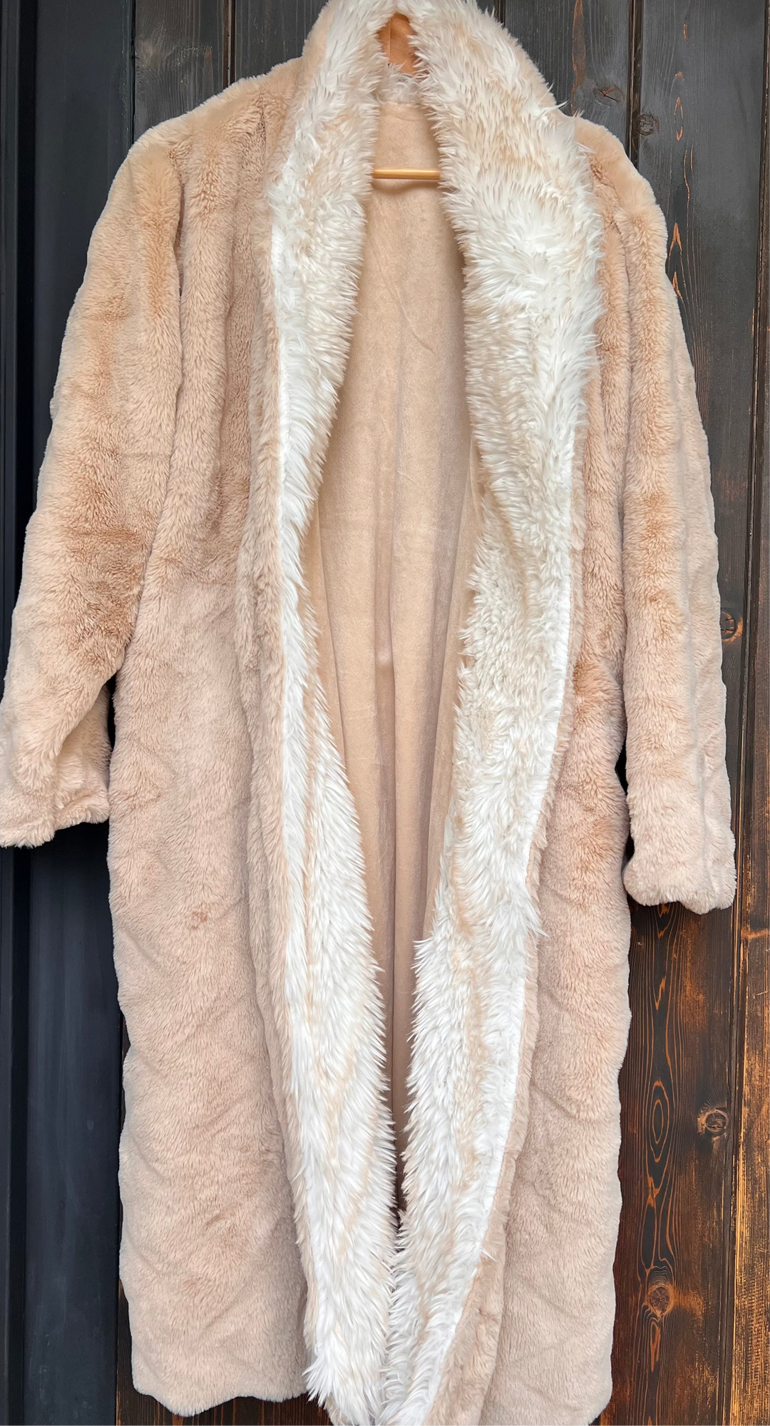 Beige fur coat with a white fur trim on a wooden hanger against a dark wooden background.