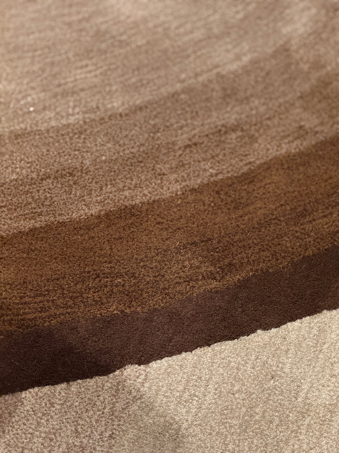 Close-up of a textured fabric or carpet sample with horizontal bands in warm beige, medium brown, and dark brown.