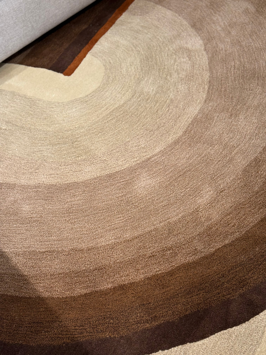 A close-up of a round area rug with concentric circular bands in beige, taupe, and dark brown, with a portion of a gray upholstered chair visible at the top left.