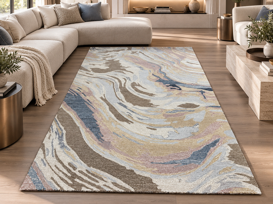A rectangular area rug with an abstract marbled pattern in beige, blue, gray, and soft pink tones, styled in a modern living room setting with a light beige sectional sofa and wood flooring.